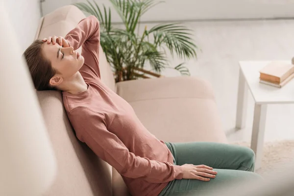 Selective focus of girl sitting on sofa with closed eyes and suffering from migraine — Stock Photo