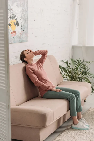 Junge Frau sitzt auf Sofa und schaut auf, während sie unter Kopfschmerzen leidet — Stockfoto