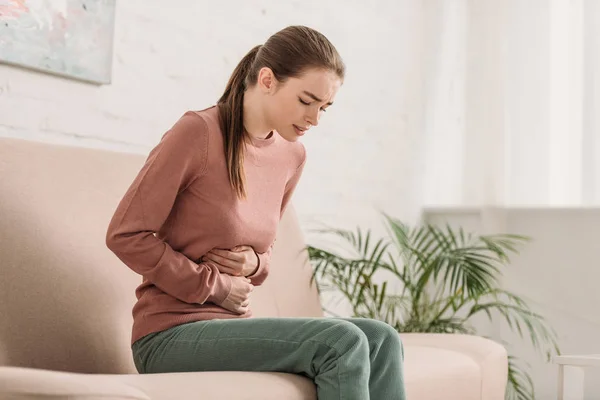 Unhappy girl sitting on sofa and suffering from stomach pain — Stock Photo