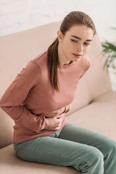 Menina franzida sentado no sofá e sofrendo de dor de estômago — Fotografia de Stock