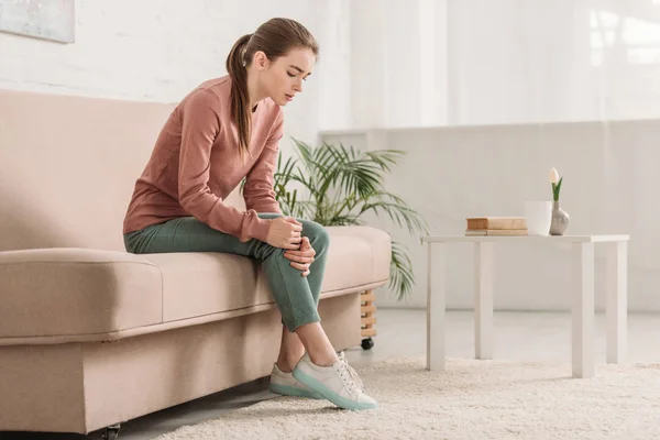 Upset girl sitting on sofa and suffering from knee pain — Stock Photo