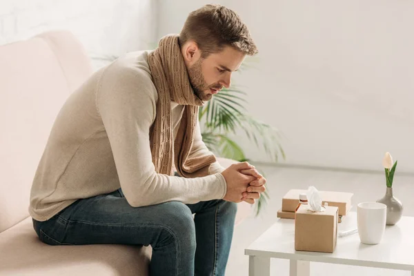 Sick man in warm scarf and sweater sitting on sofa near table with medicines — Stock Photo