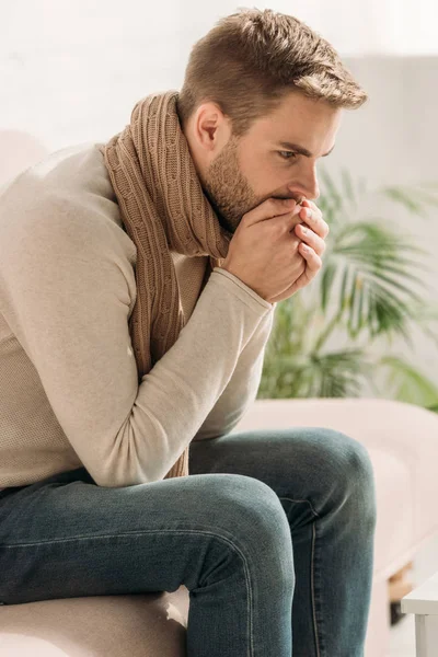 Hombre enfermo en bufanda caliente tosiendo en las manos dobladas mientras está sentado en el sofá - foto de stock