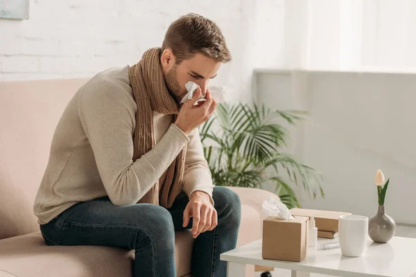Ill man sneezing in napkin while sitting on sofa near table — Stock Photo