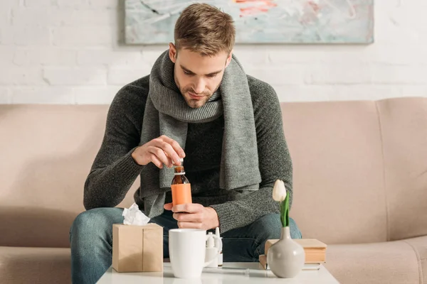 Kranker Mann sitzt auf Sofa und öffnet Flasche mit Hustensaft — Stockfoto