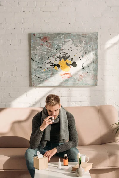 Ill man sitting on sofa near table with medicines and using nasal spray — Stock Photo