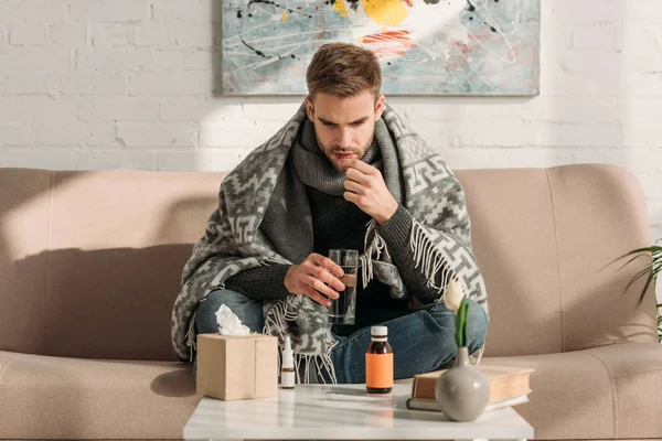 Uomo malato, avvolto in una coperta, seduto sul divano e che prende medicine — Foto stock