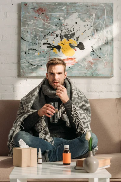 Sick man, wrapped in blanket, sitting on sofa and taking medicines — Stock Photo