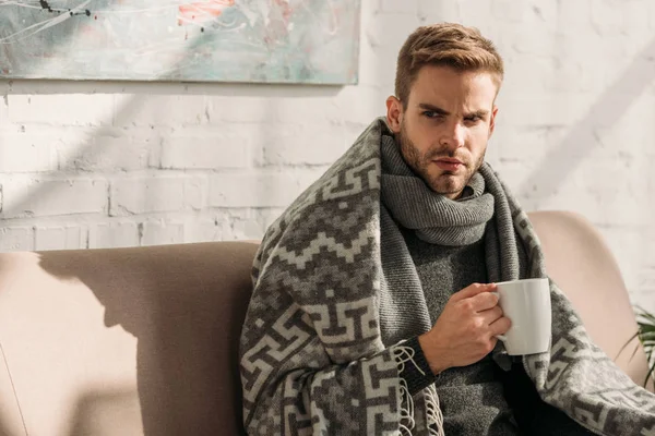 Enfermo, hombre infeliz, envuelto en manta, mirando hacia otro lado mientras sostiene la taza de bebida caliente - foto de stock