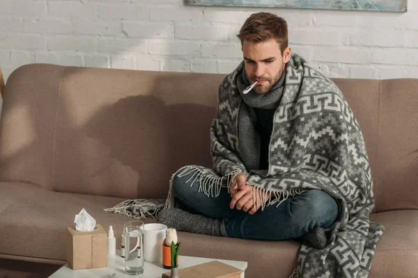 Hombre enfermo y molesto midiendo la temperatura mientras está sentado en el sofá cerca de la mesa con medicamentos - foto de stock