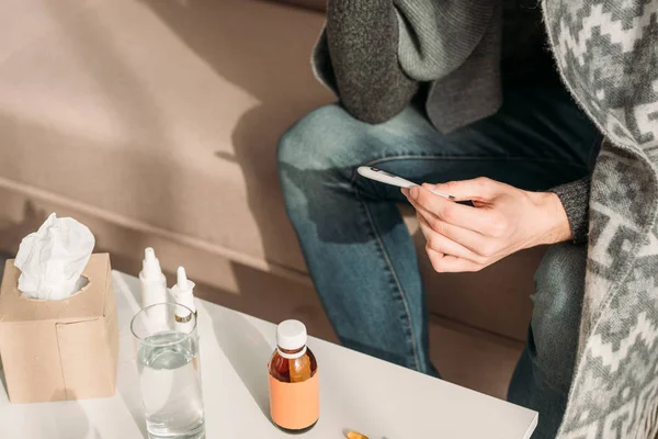 Vista recortada del hombre enfermo sentado cerca de la mesa con medicamentos y termómetro de retención - foto de stock