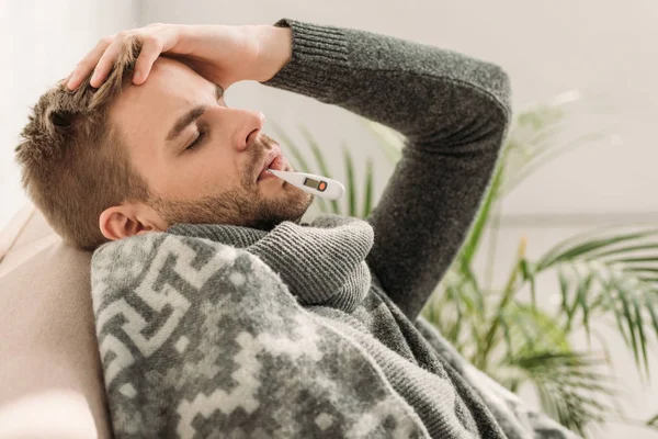 Malato, avvolto in una coperta, seduto sul divano con gli occhi chiusi e la temperatura di misura — Foto stock