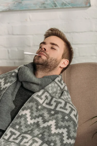 Enfermo, envuelto en manta, sentado en un sofá con los ojos cerrados y midiendo la temperatura - foto de stock