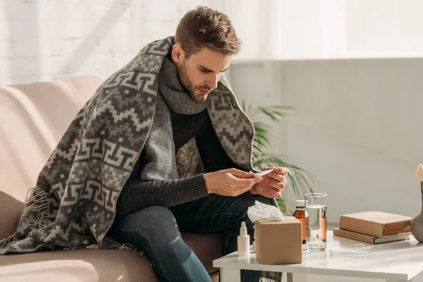 Hombre enfermo, envuelto en manta, sentado en el sofá y mirando el termómetro - foto de stock