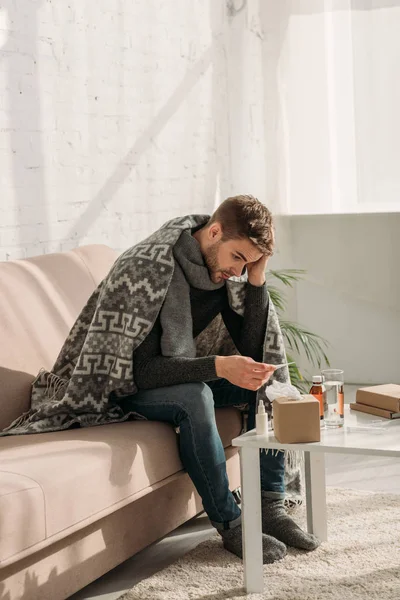 Больной человек, завернутый в одеяло, сидит на диване и смотрит на термометр — стоковое фото