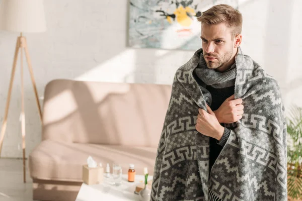 Sick, unhappy man, wrapped in blanket, looking away while standing in room — Stock Photo