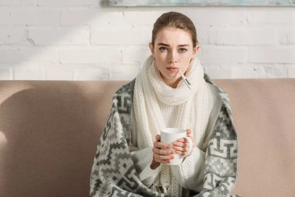 Fille malade regardant la caméra tout en mesurant la température et tenant tasse de boisson chauffante — Photo de stock