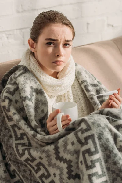 Krankes Mädchen, in Decke gehüllt, blickt in die Kamera, während es Thermometer und Becher mit wärmendem Getränk in der Hand hält — Stockfoto
