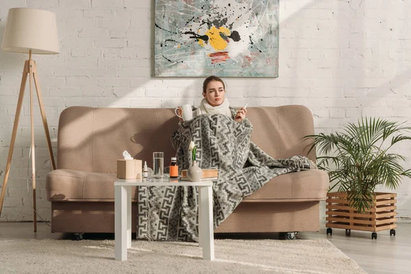 Kranke Frau hüllt sich in Decke, während sie auf Sofa sitzt und Thermometer und Becher mit wärmendem Getränk in der Hand hält — Stockfoto