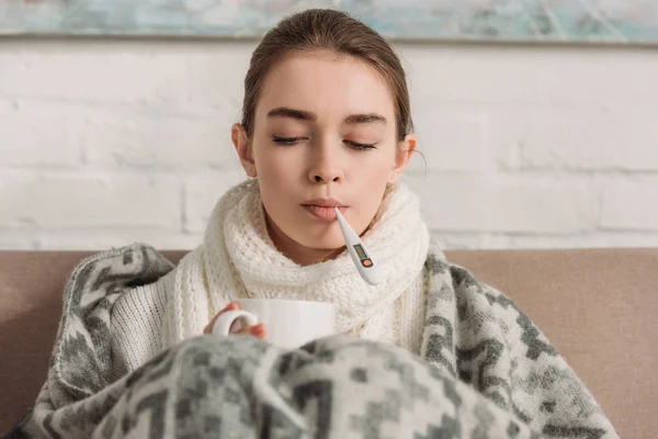 Femme malade mesurant la température tout en tenant tasse de boisson chauffante — Photo de stock