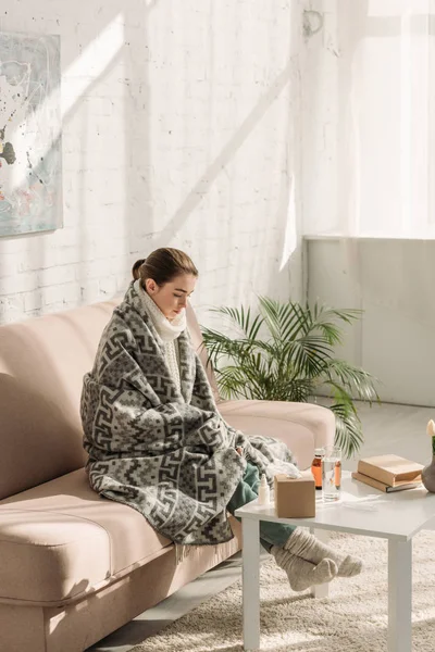 Fille malade, enveloppé dans une couverture, assis sur le canapé près de la table avec des livres et des médicaments — Photo de stock