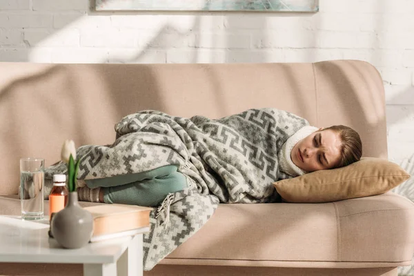 Femme malade couchée sur le canapé sous couverture près de la table avec des médicaments — Photo de stock