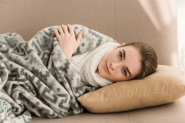 Mujer enferma y positiva tumbada en el sofá debajo de la manta y mirando a la cámara - foto de stock