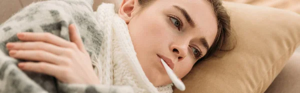 Panoramic shot of sick woman lying on sofa under blanket and measuring temperature — Stock Photo