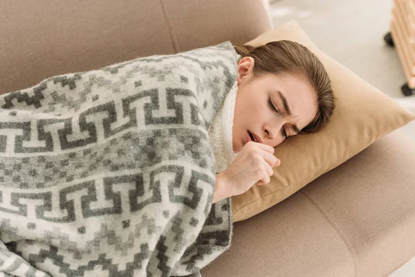 Chica enferma tosiendo mientras está acostada en el sofá debajo de la manta - foto de stock