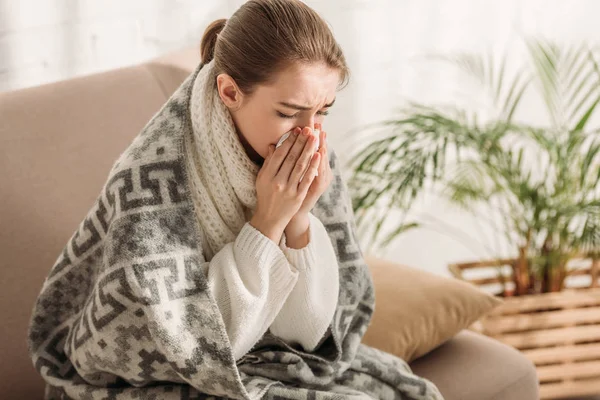 Sick woman sneezing in napkin while sitting on sofa and wrapping in blanket — Stock Photo