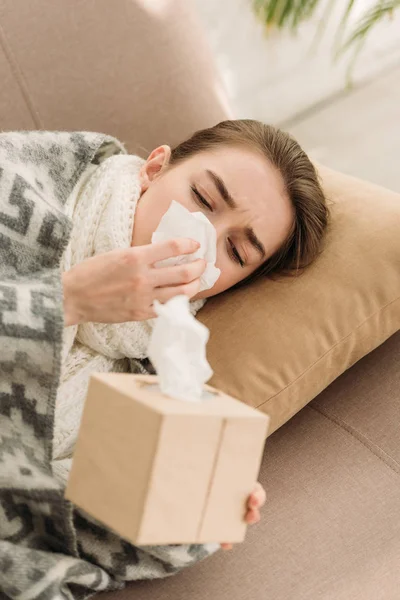 Sick girl, covered with blanket, lying on sofa and sneezing in napkin — Stock Photo