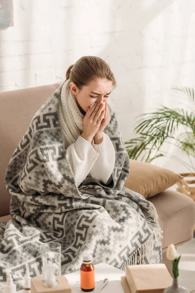 Femme malade, enveloppée dans une couverture, assise sur un canapé et éternuant dans une serviette — Photo de stock