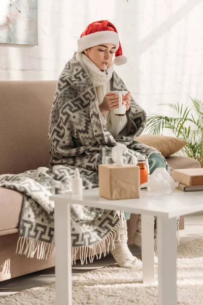 Sick woman in santa hat, wrapped in blanket, measuring temperature and holding cup of warming drink — Stock Photo