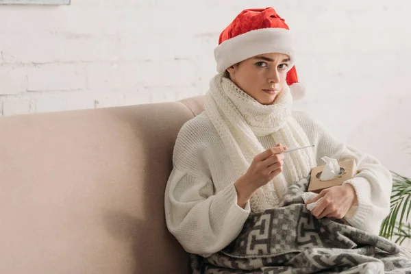 Kranke Frau mit Weihnachtsmütze hält Thermometer in der Hand und blickt in die Kamera — Stockfoto