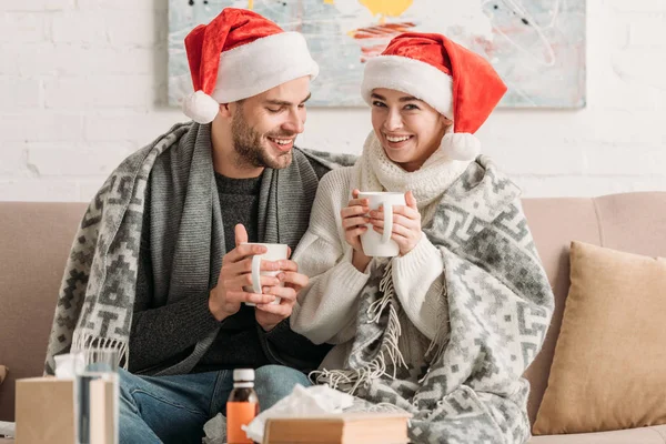 Diseased, smiling couple in santa hats, covered with blanket, holding cups of warming drink while sitting on sofa — Stock Photo