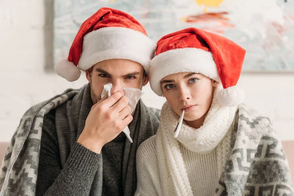 Diseased couple wearing santa hats and covered with blanket looking at camera — Stock Photo
