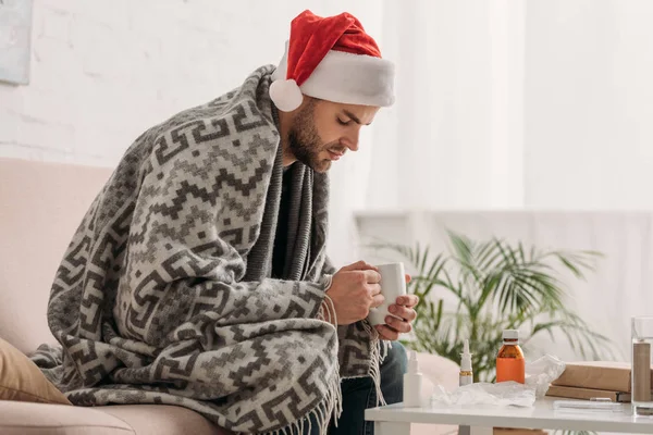 Malade au chapeau de Père Noël, enveloppé dans une couverture, assis près de la table avec des médicaments et tenant une tasse de boisson chauffante — Photo de stock