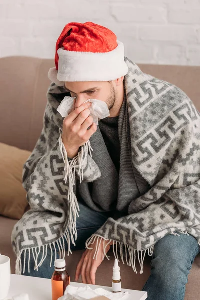 Sick man sitting in santa hat, wrapped in blanket, sitting in sofa and sneezing in napkin — Stock Photo