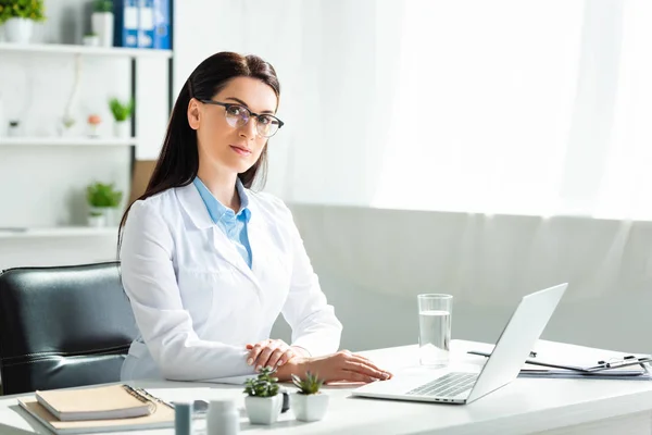 Serious doctor sitting in clinic office with laptop — Stock Photo