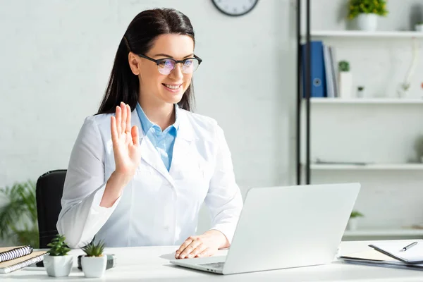 Médico sorrindo acenando e tendo consulta on-line com o paciente no laptop no escritório da clínica — Fotografia de Stock