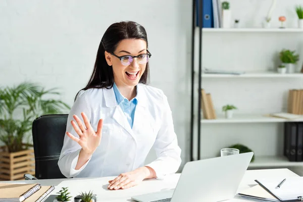 Excited doctor waving and having online consultation with patient on laptop in clinic office — Stock Photo