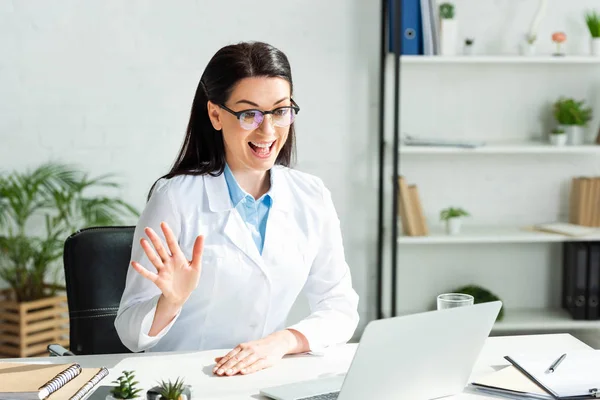 Excitado médico saludando y teniendo consulta en línea con el paciente en el ordenador portátil en la clínica - foto de stock