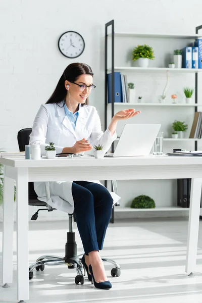 Улыбающаяся женщина врач, имеющая онлайн консультации с пациентом на ноутбуке в офисе — стоковое фото