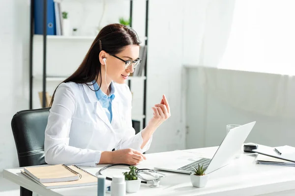 Female positive doctor in earphones having online consultation with patient on laptop in clinic office — Stock Photo