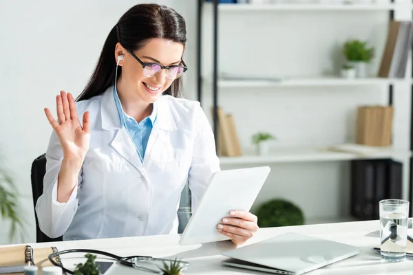Positive doctor waving and having online consultation on digital tablet in clinic office with laptop — Stock Photo