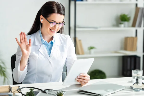 Doctor positivo saludando y teniendo consulta en línea en la tableta digital en la oficina clínica con el ordenador portátil - foto de stock