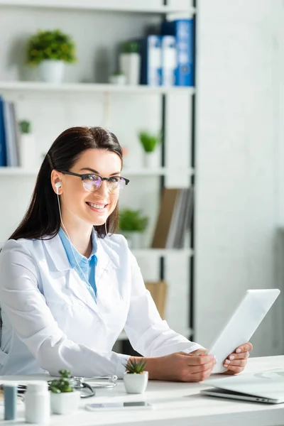 Позитивний лікар, який має онлайн-консультацію на цифровому планшеті в клінічному офісі з ноутбуком — стокове фото