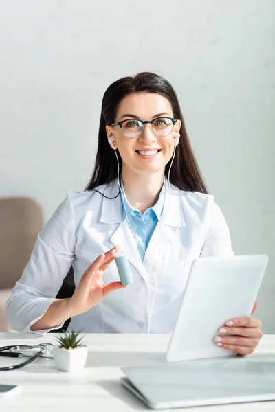 Médico sonriente en auriculares sosteniendo pastillas y tableta digital en la clínica - foto de stock