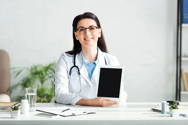Atractivo médico sonriente mostrando tableta digital con pantalla en blanco en la oficina de la clínica - foto de stock