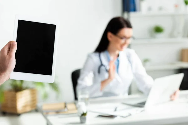 Обрізаний вид людини, що тримає цифровий планшет з порожнім екраном в клінічному кабінеті з лікарем, який має онлайн-консультацію на ноутбуці — стокове фото